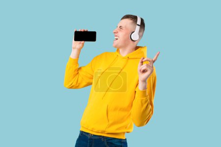 Téléchargez les photos : Portrait d'un caucasien ravi chantant et utilisant le téléphone cellulaire comme microphone, portant un casque sans fil. Jeune homme s'amusant sur fond bleu studio - en image libre de droit