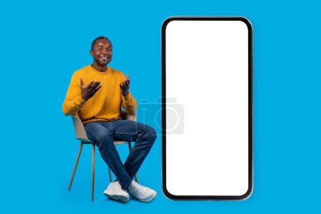 Photo for Happy handsome middle aged black man in casual sitting on chair by big cell phone with white empty screen, smiling and gesturing, mockup for online offer, blue studio background, full length - Royalty Free Image