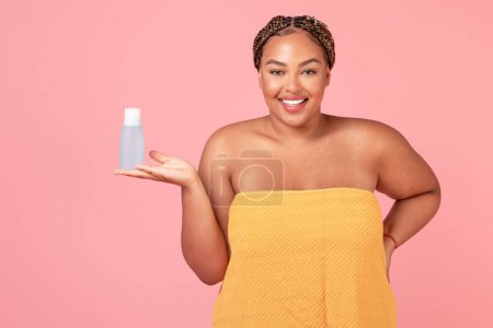Téléchargez les photos : Corps positif femme afro-américaine tenant bouteille d'eau micellaire, la publicité de nouveaux produits cosmétiques, posant sur fond rose, plan studio - en image libre de droit