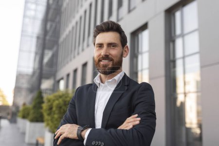 Photo for Confident glad young european male ceo manager with beard crossed his arms over chest, looks at empty space near building on city street, close up. Leader, finance, startup, modern business and boss - Royalty Free Image