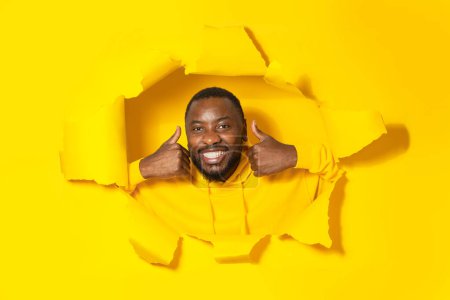 Photo for Joyful black man gesturing thumbs up and smiling at camera through hole in torn yellow paper, approving great offer, posing in studio. I like it concept - Royalty Free Image