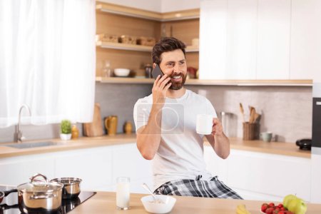 Téléchargez les photos : Heureux homme caucasien adulte avec barbe en t-shirt blanc boit tasse de café, appels par téléphone dans l'intérieur de la cuisine moderne. Potins, bonnes nouvelles, communication à distance, bonjour et petit déjeuner à la maison - en image libre de droit