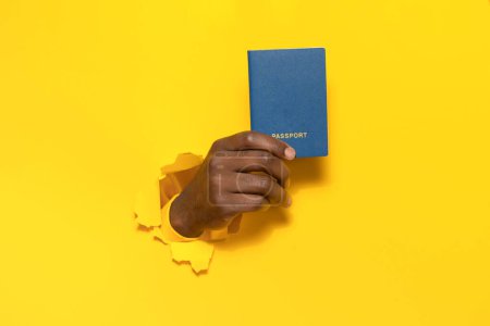 Foto de Viajero afroamericano turista mano masculina sosteniendo pasaporte de un agujero roto en fondo de pared de papel amarillo, espacio libre de copia, primer plano - Imagen libre de derechos