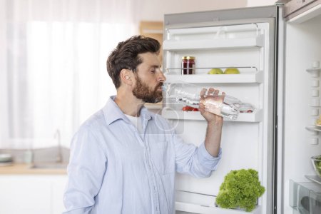Photo for Adult thirst caucasian man with beard in white shirt opens refrigerator door, drinks water from bottle, enjoy fresh in kitchen interior, free space. Health care, proper nutrition, aqua balance at home - Royalty Free Image
