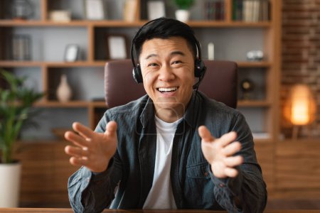 Photo for Cheerful adult japanese male with headphones gesticulates hands, looks at computer webcam, has business video call in home office interior, close up. Work remotely, meeting webinar and new normal - Royalty Free Image