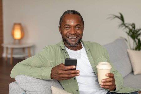 Photo for Coffee break with gadget. Happy middle aged african american man on sofa watch video on smartphone, enjoy cup of drink takeaway in living room interior. Chat in social networks, app and rest at home - Royalty Free Image