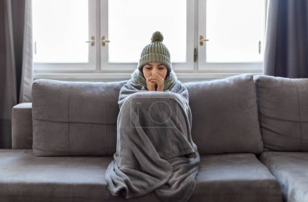 Photo for Cold Home. Young Woman Covered With Blanket Freezing On Couch In Living Room, Millennial Female Wearing Knitted Scarf And Hat Indoors Warming Hands With Her Breath, Suffering Heating Problems - Royalty Free Image