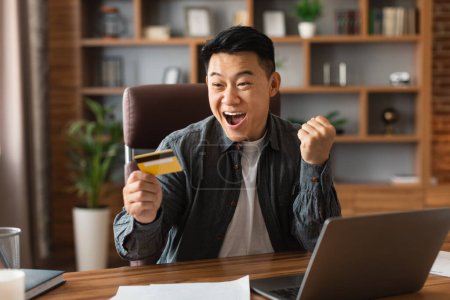Foto de Satisfecho sorprendido adulto adulto japonés hombre de negocios con la boca abierta celebrar tarjeta de crédito, hacer gesto de victoria en el interior de la oficina en casa. Victoria, trabajo exitoso, negocios a distancia finanzas y emociones - Imagen libre de derechos