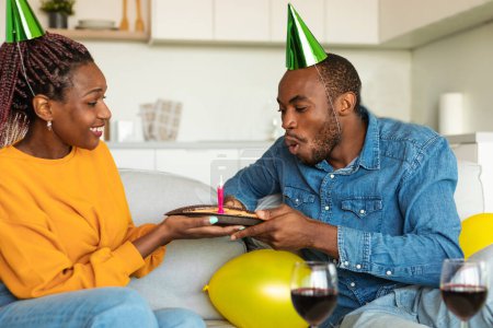 Foto de Amar pareja afroamericana en sombreros de fiesta celebrando cumpleaños, hombre soplando vela en pastel festivo, sentado en el sofá en casa. Jóvenes cónyuges negros que tienen fiesta doméstica b-day - Imagen libre de derechos