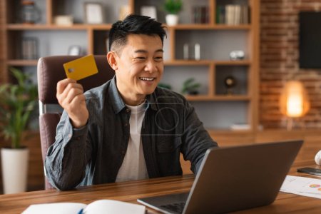 Sonriendo hombre asiático adulto mostrar tarjeta de crédito, trabajar en la computadora, utilizar la banca financiera para las compras en línea en el interior de la oficina en casa. Recomendación y asesoramiento para la inversión, el trabajo, el negocio remoto