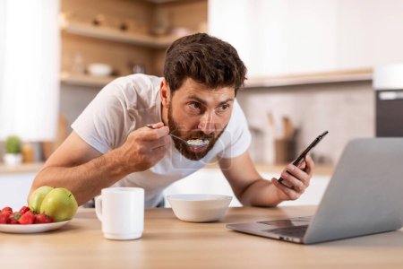 Téléchargez les photos : Homme caucasien adulte sérieux occupé avec barbe en t-shirt mange du porridge, utilise un smartphone, regarde des vidéos sur ordinateur portable à l'intérieur de la cuisine. App pour le travail et les affaires, bonjour et petit déjeuner, nouvelles à la maison - en image libre de droit