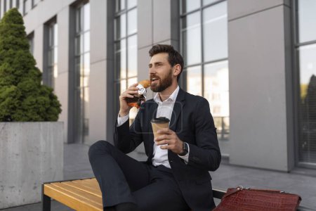 Photo for Happy hangry young european male manager with beard in suit with cup of coffee takeaway eats croissant, sits on bench near building in city. Food, lunch outdoor, break from work and rest at free time - Royalty Free Image