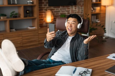 Foto de Satisfecho adulto emocionado hombre asiático en auriculares inalámbricos mirando webcam smartphone, tiene videollamada en la mesa en el interior de la oficina en casa. El descanso de los negocios, ver vídeo en el dispositivo durante el descanso, covid-19 - Imagen libre de derechos