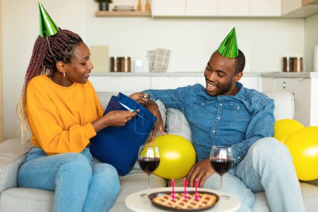 Photo for Happy birthday my love. Excited black woman giving gift to her husband, young couple sitting on couch at home. Loving wife opening shopping bag, showing present to man - Royalty Free Image