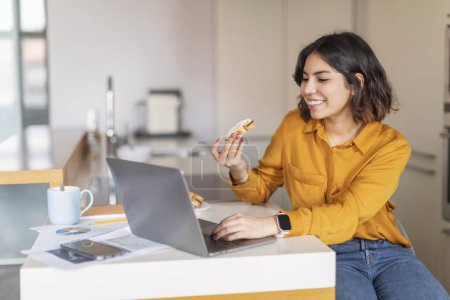 Foto de Mujer árabe joven sonriente comiendo bocadillo y usando el ordenador portátil en la cocina, freelancer femenina de Oriente Medio feliz disfrutando pedazo de pastel y viendo webinar en línea en la computadora en casa, espacio de copia - Imagen libre de derechos