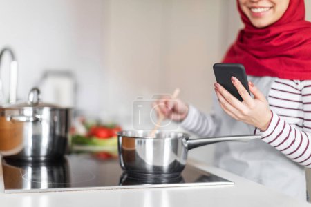 Téléchargez les photos : Coupé de femme en hijab et tenue décontractée cuisine à Kicthen et en utilisant un smartphone, dame musulmane méconnaissable tenant téléphone cellulaire, en utilisant l'application de recette ou blog alimentaire en ligne, copier l'espace - en image libre de droit