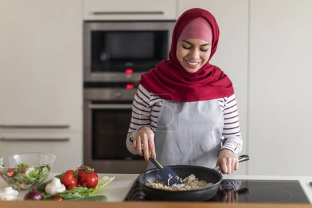 Foto de Mujer joven bastante musulmana en hijab con delantal prepara comida saludable para su familia en la cocina moderna en casa, cocinar deliciosa cena llena de verduras, espacio para copiar. Concepto de cocina - Imagen libre de derechos