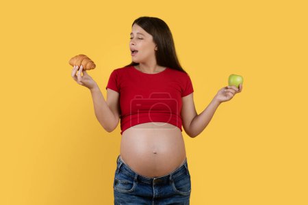 Foto de Embarazo Nutrición. Mujer joven embarazada eligiendo Croissant en lugar de manzana verde, hermosa mujer embarazada tomando la decisión entre la comida sana y poco saludable mientras está de pie sobre el fondo amarillo - Imagen libre de derechos