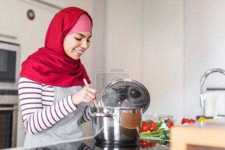 Téléchargez les photos : Joyeux jeune femme arabe portant hijab, tablier et tenue décontractée confortable cuisine femme au foyer sur cuisinière électrique à la maison, mélangeant la nourriture dans la casserole et souriant, copier l'espace. Cuisine, concept de style de vie domestique - en image libre de droit