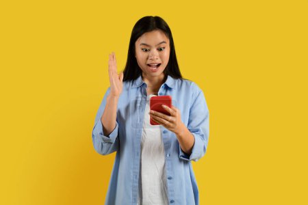 Photo for Cheerful surprised millennial korean female with down syndrome in casual with open mouth reads message on phone isolated on yellow background, studio. Shocking ad and offer, victory, success emotions - Royalty Free Image