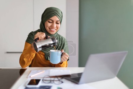 Téléchargez les photos : Sourire musulmane indépendante assise au bureau avec ordinateur portable et verser du café de Prench Press, jeune femme arabe dans le hijab se préparer avant de travailler en ligne avec ordinateur, Copier l'espace - en image libre de droit