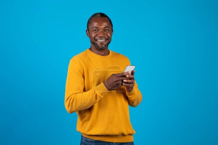 Téléchargez les photos : Photo studio d'un homme afro-américain adulte joyeux en tenue décontractée utilisant un tout nouveau téléphone cellulaire isolé sur fond bleu, jouant ou commerçant sur Internet, regardant la caméra et souriant, espace de copie - en image libre de droit