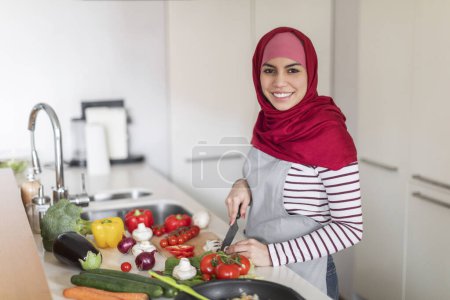 Téléchargez les photos : Joyeux jeune belle femme portant foulard femme musulmane debout à côté de la table de cuisine pleine d'aliments biologiques, couper des légumes pour un délicieux dîner sain, sourire à la caméra, copier l'espace - en image libre de droit