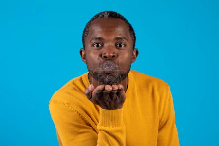 Téléchargez les photos : Portrait photo de l'homme noir afro-américain d'âge moyen en jaune doux romantique envoyer baiser d'air à la caméra isolé fond bleu studo. Amour, sentiments, flirt, gestes humains concept - en image libre de droit