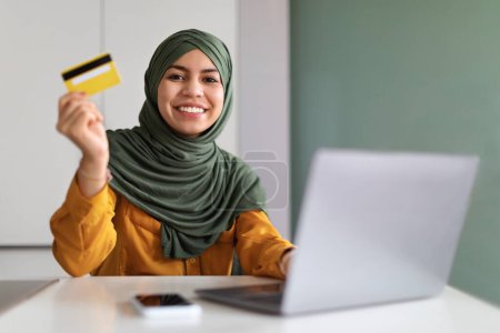 Photo for E-Commerce Concept. Smiling Muslim Lady Holding Creadit Card And Using Laptop While Sitting At Desk In Home Office, Young Middle Eastern Woman In Hijab Enjoying Purchasing From Internet, Free Space - Royalty Free Image