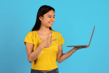 Téléchargez les photos : Annonce et offre, vente énorme. Heureuse jeune étudiante asiatique en t-shirt jaune pointeur doigt à l'écran de l'ordinateur isolé sur fond bleu. Recommandation, conseils d'un professionnel, dispositif de travail, étude - en image libre de droit