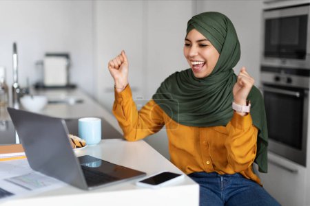Téléchargez les photos : Super nouvelle. Jeune femme musulmane heureuse dans le Hijab célébrant le succès avec l'ordinateur portable tout en travaillant dans la cuisine à la maison, joyeuse femme islamique regardant l'écran d'ordinateur, levant les poings et exclure avec joie - en image libre de droit