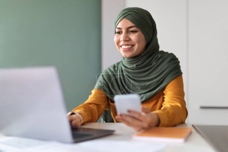 Photo for Portrait Of Young Arab Woman In Hijab Using Smartphone And Laptop While Working At Home Office, Millennial Muslim Woman Wearing Headscarf Sitting At Desk And Typing On Computer, Enjoying Online Job - Royalty Free Image