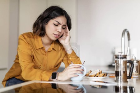 Photo for Depression Concept. Upset Arab Woman Sitting At Kitchen Counter At Home, Closeup Shot Of Young Depressed Middle Eastern Female Touching Head, Feeling Unwell While Drinking Morning Coffee, Free Space - Royalty Free Image
