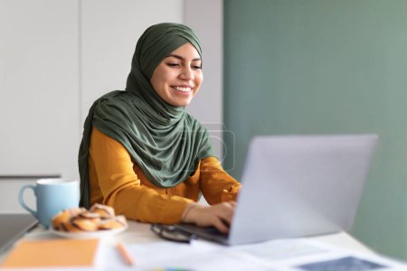 Foto de Concepto de Educación en Línea. Sonriente joven musulmana en Hijab usando el ordenador portátil en casa, feliz señora de Oriente Medio sentado en el escritorio con la computadora y escribiendo en el teclado, disfrutando de la educación a distancia - Imagen libre de derechos