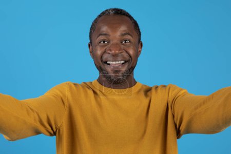 Photo for Positive happy handsome mature african american man in yellow taking selfie, cheerfully smiling at camera, showing white teeth, isolated on blue studio background, headshot - Royalty Free Image
