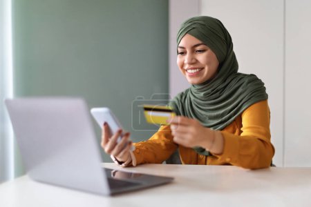 Téléchargez les photos : Paiements en ligne. Femme musulmane utilisant smartphone et carte de crédit tout en étant assis au bureau avec ordinateur portable au bureau à la maison, souriant dame islamique dans hijab payer des factures sur Internet ou transférer de l'argent via l'application - en image libre de droit