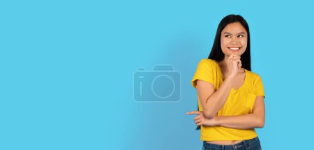 Foto de Me alegra bastante millennial estudiante de mujer japonesa en camiseta amarilla mira el espacio vacío y piensa en el nuevo orden, aislado en el fondo azul, estudio, panorama. Gran anuncio y oferta, venta, emociones humanas - Imagen libre de derechos