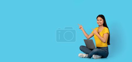 Téléchargez les photos : Heureuse étudiante blogueuse asiatique millénaire est assise sur le sol avec un ordinateur pointant du doigt l'espace vide, isolée sur fond bleu, panorama, studio. Gadget pour le travail et l'étude, la vente, l'annonce et l'offre - en image libre de droit