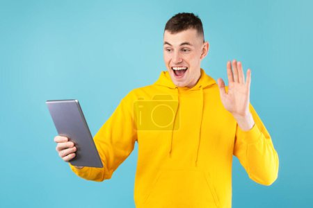 Photo for Video call. Excited caucasian man chatting online via digital tablet and waving hand to webcam, standing over blue background. Modern remote communication - Royalty Free Image