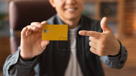 Téléchargez les photos : Heureux homme asiatique d'âge moyen pointant du doigt la carte de crédit à l'intérieur du bureau à la maison, recadré. Recommandation d'homme d'affaires et conseils pour l'investissement, le travail, les affaires et les finances à distance pendant covid-19 - en image libre de droit