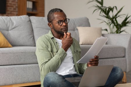 Photo for Concentrated pensive middle aged african american guy in glasses with laptop reads document in living room interior. Work and business, new normal, accounting and data analysis, startup bookkeeping - Royalty Free Image