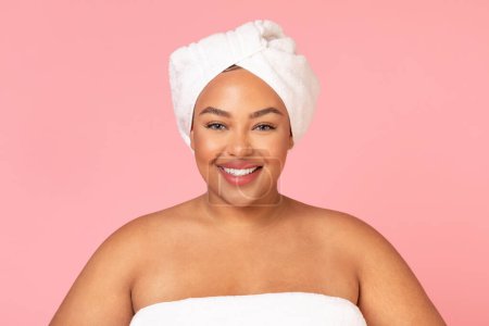 Téléchargez les photos : Portrait de beauté du corps positif femme noire debout enveloppé dans une serviette sur fond rose et souriant à la caméra. Belle femme obèse à la peau lisse posant après le bain - en image libre de droit