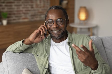 Téléchargez les photos : Portrait de joyeux homme afro-américain d'âge moyen dans des lunettes appels par smartphone, gesticule, parle dans le salon intérieur, gros plan. Dispositif de communication, commérages et bonnes nouvelles à la maison - en image libre de droit