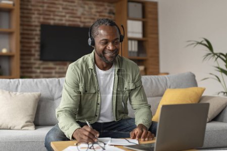 Photo for Smiling middle aged african american guy in headphones looks at laptop, makes notes in living room interior. Video call, online lesson, customer support, work and business remote, new normal at home - Royalty Free Image