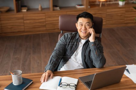 Téléchargez les photos : Heureux confiant succès adulte asiatique homme d'affaires appelez par téléphone, asseyez-vous à table avec ordinateur à l'intérieur du bureau à la maison. Télécommande d'entreprise moderne, financement d'investissement et dispositif de communication, covid-19 - en image libre de droit