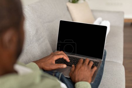 Téléchargez les photos : Occupé homme afro-américain d'âge moyen sur canapé regarder la vidéo, dactylographier sur ordinateur portable avec écran blanc à temps libre dans le salon intérieur. Chat sur gadget, application, site Web pour le travail, l'étude et les affaires à la maison - en image libre de droit
