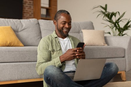 Photo for App for work, business remote. Happy middle aged african american male with laptop typing on phone, chatting in social networks in living room interior. Surfing in internet at home, social distance - Royalty Free Image