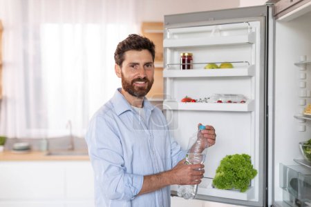 Photo for Cheerful mature caucasian male with beard in shirt opens refrigerator door, takes bottle of water in kitchen interior. Aqua balance, diet, health care and lifestyle at home due covid-19 quarantine - Royalty Free Image
