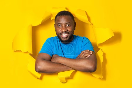 Photo for Portrait of positive black man posing with folded arms in torn paper hole, looking and smiling at camera through breakthrough of yellow background. - Royalty Free Image