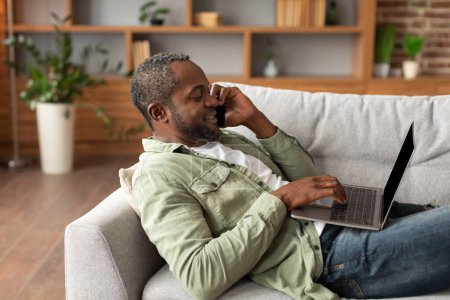 Téléchargez les photos : Heureux occupé mi-âge afro-américain mâle tapant sur ordinateur portable, appelant par smartphone, choisissant l'ordre, bavardant dans les médias sociaux dans le salon intérieur. Travail et affaires, surf sur internet à la maison - en image libre de droit